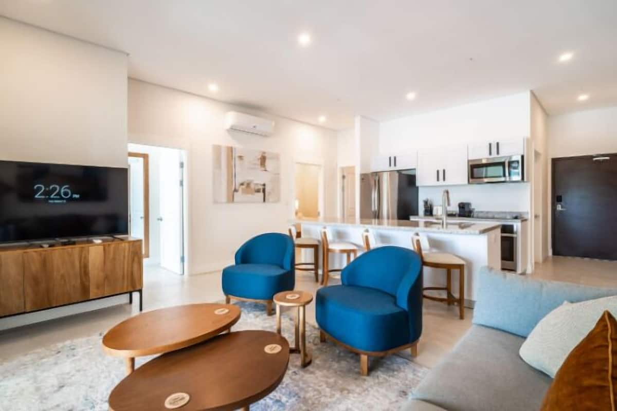 A modern living room with a TV, blue armchairs, a sofa, and a kitchen in the background.