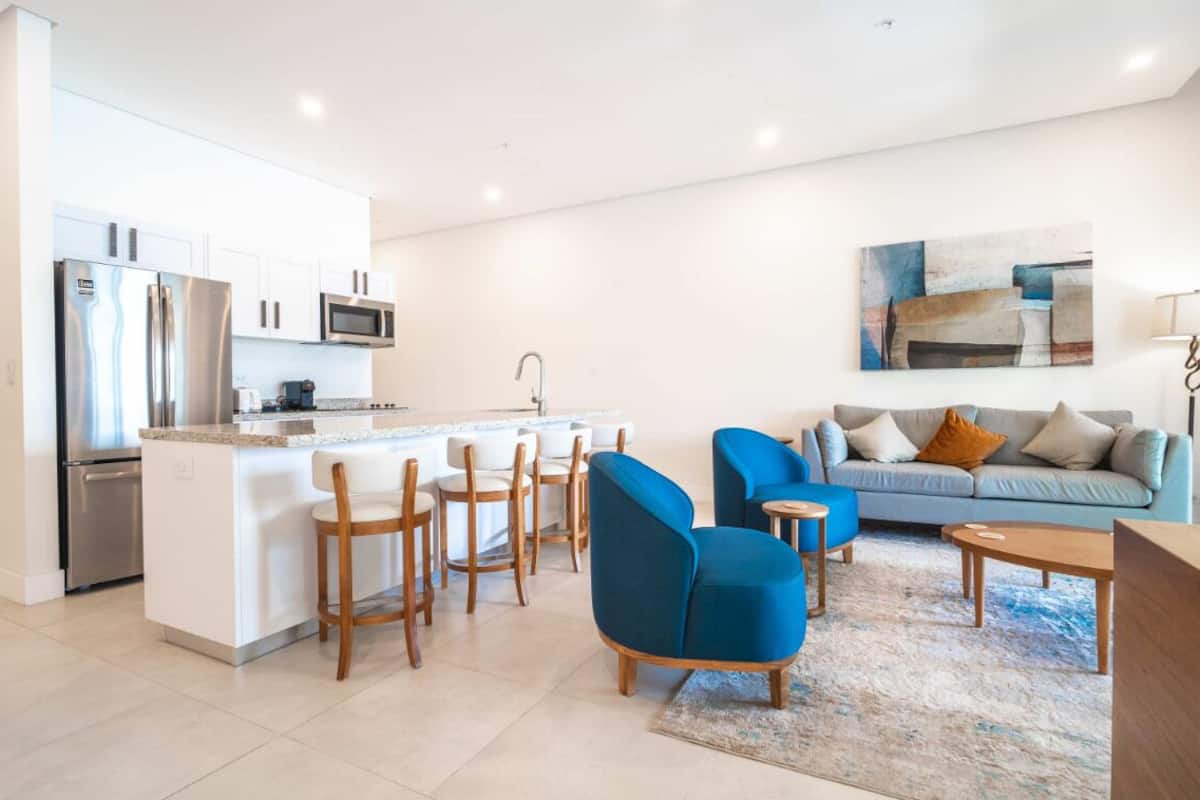 A modern kitchen with a central island, bar stools, a dining area with a blue sofa, and a living room with a light blue sofa and a painting on the wall.