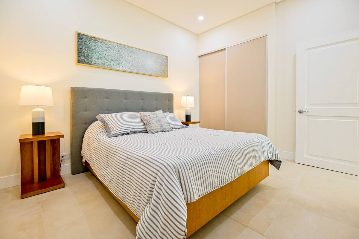 A neatly arranged bedroom with a bed, bedside lamps, a headboard, a painting, and a wardrobe.