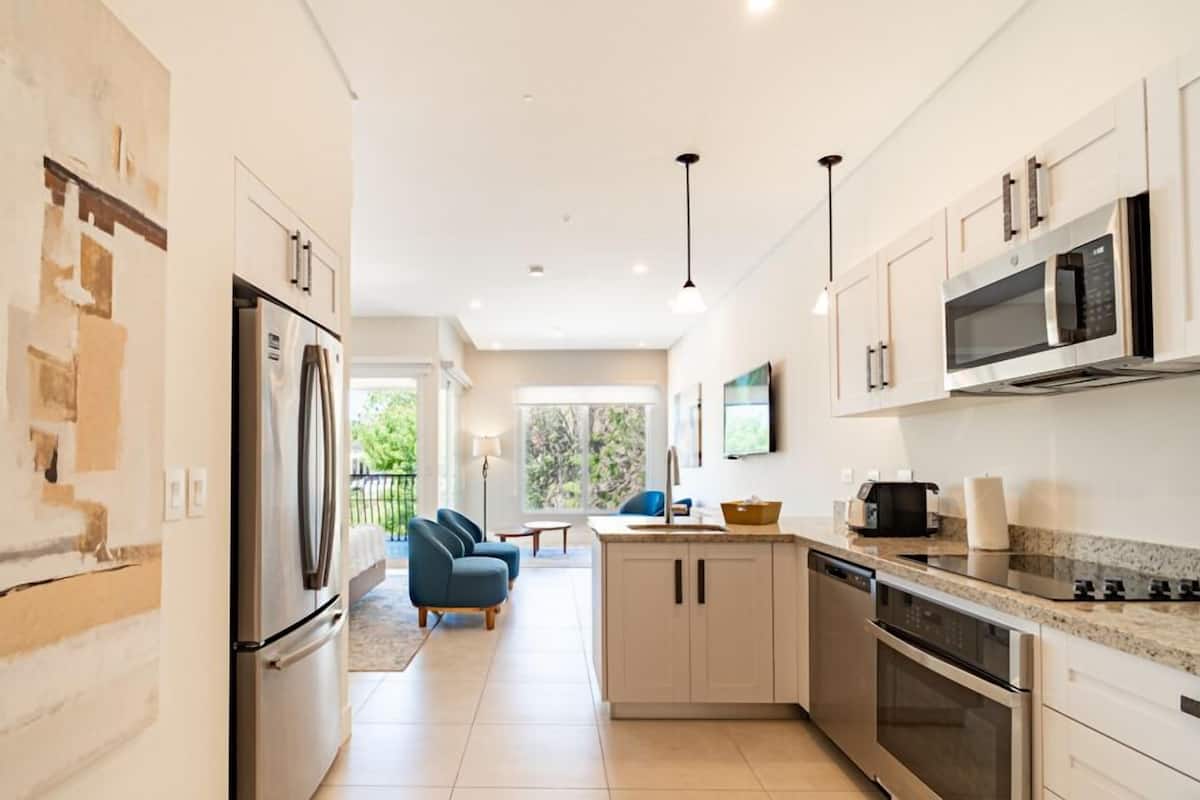 A modern kitchen with a refrigerator, oven, microwave, and dishwasher. There is a dining area with a table and chairs, and a view of greenery outside.