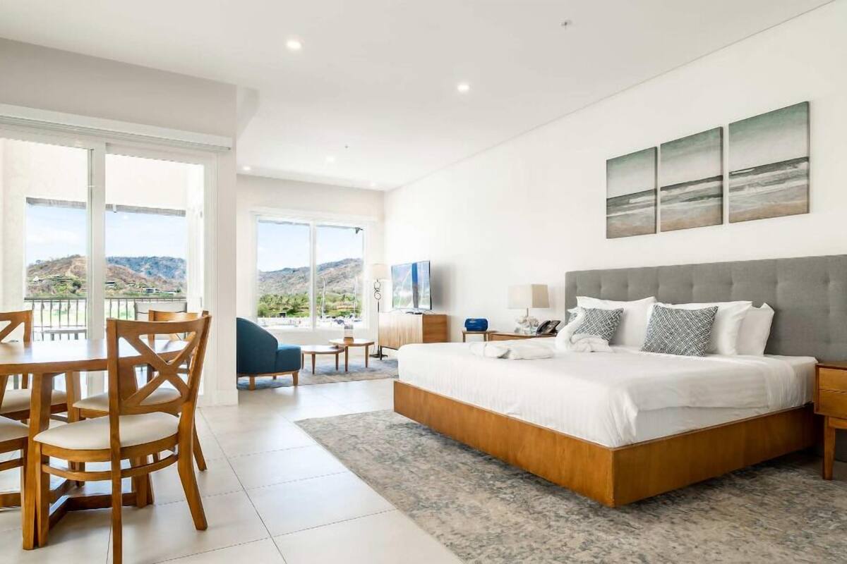 A spacious bedroom with a large bed, a desk, and chairs, and a balcony with a mountain view.