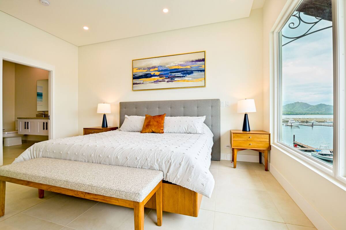 A spacious bedroom with a large bed, a bench, and a view of a marina through a large window.