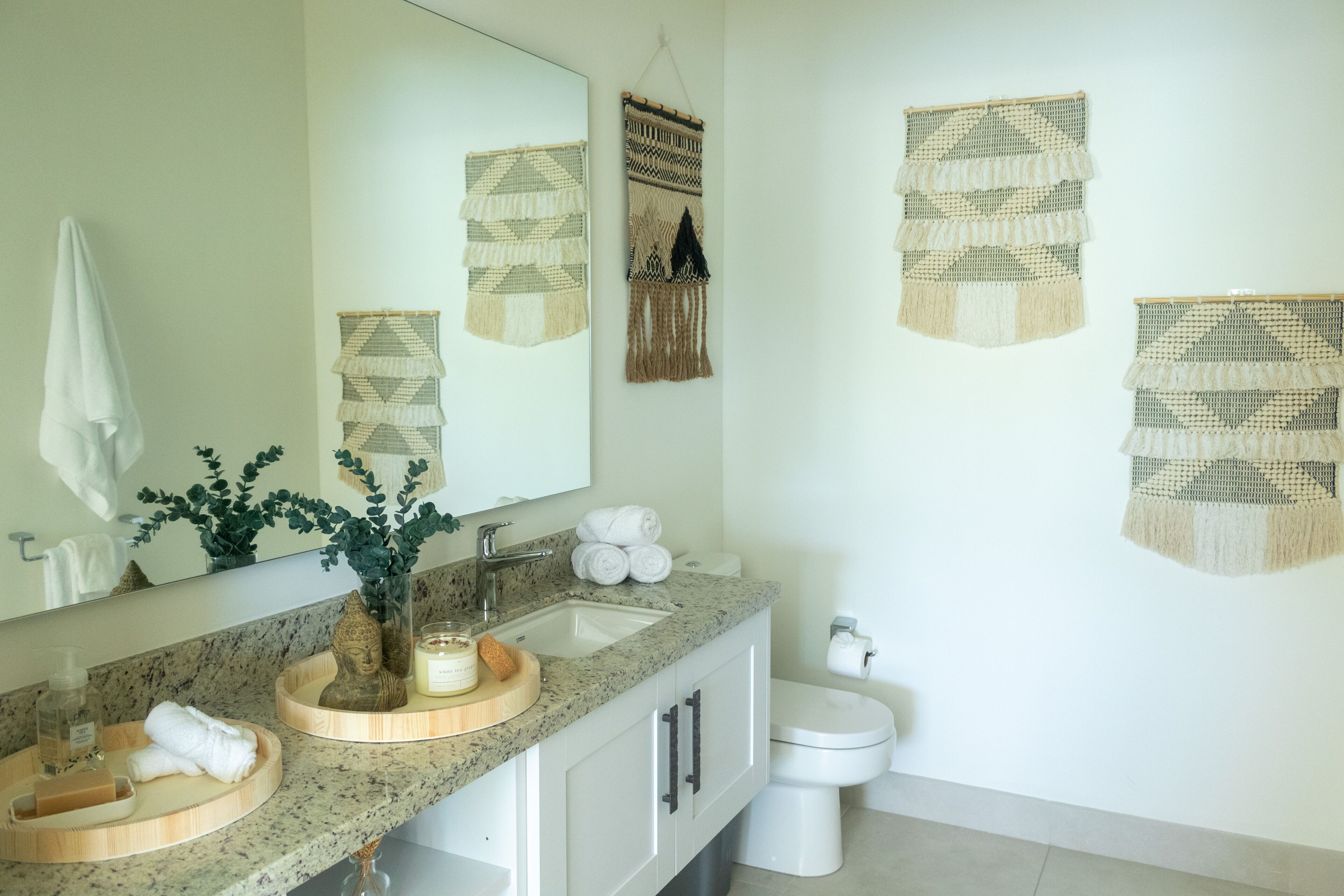 A bathroom with a granite countertop, a sink, a mirror, a toilet, and decorative wall hangings.