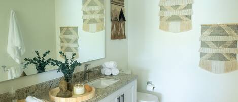 A bathroom with a granite countertop, a sink, a mirror, a toilet, and decorative wall hangings.