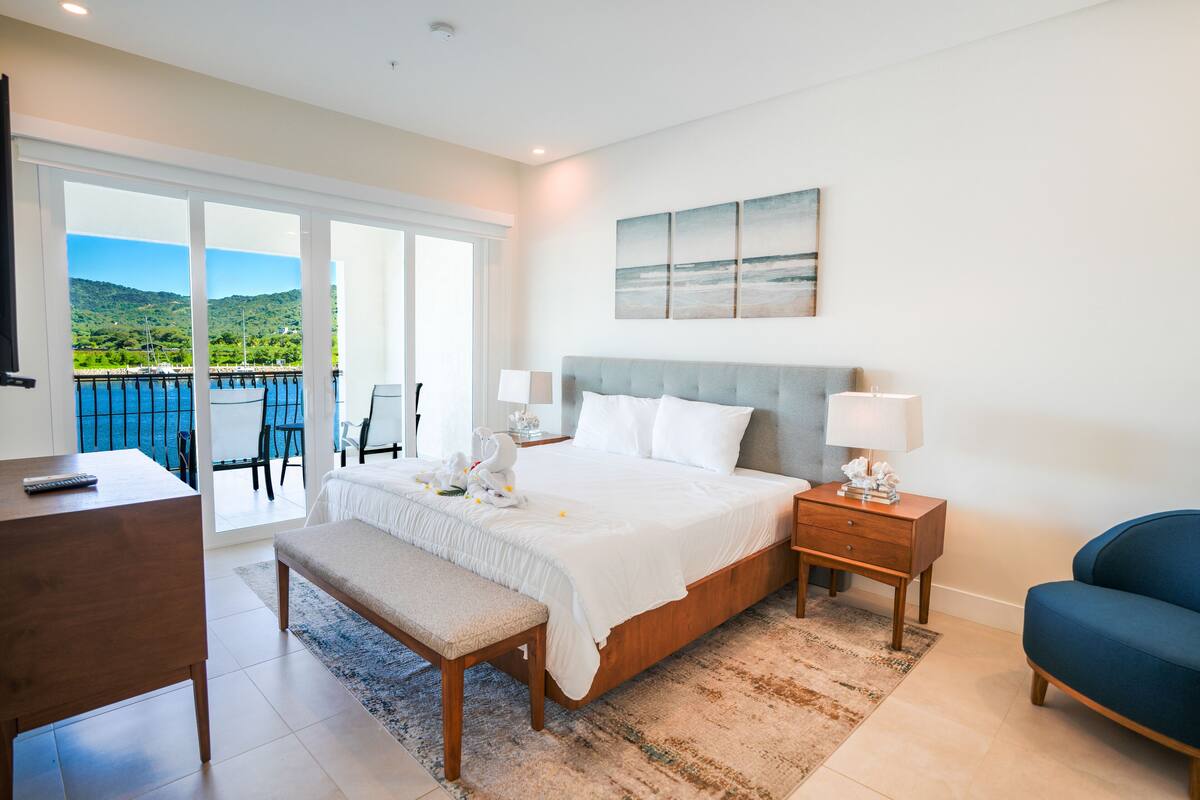 A bedroom with a large bed, a bench, a desk, a chair, a nightstand, and a view of a balcony with a scenic landscape.