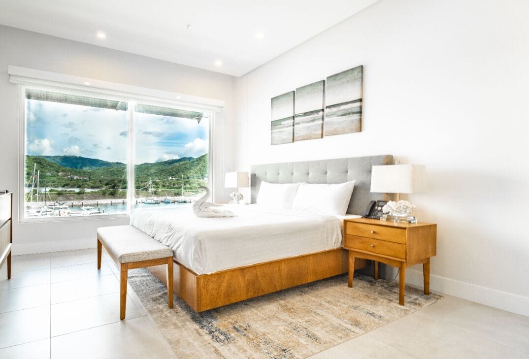A bedroom with a large window, a bed, a bedside table, and a nightstand.