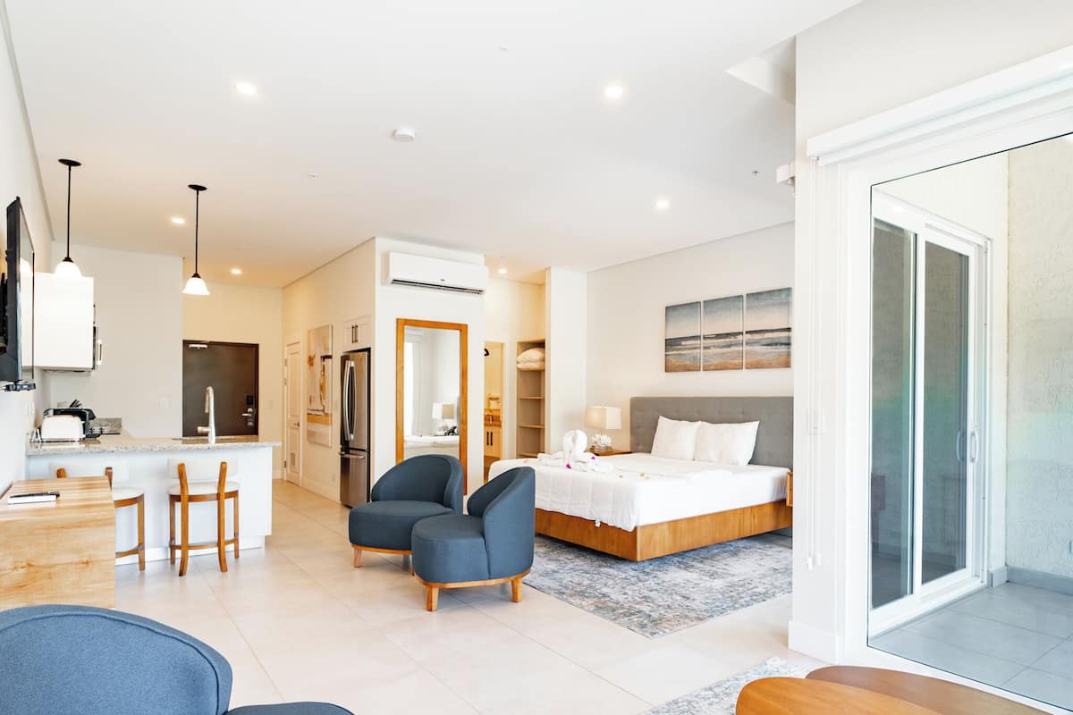 A modern hotel room with a large bed, a sitting area with two chairs, a kitchen area with a dining table and chairs, and a sliding glass door to a balcony.