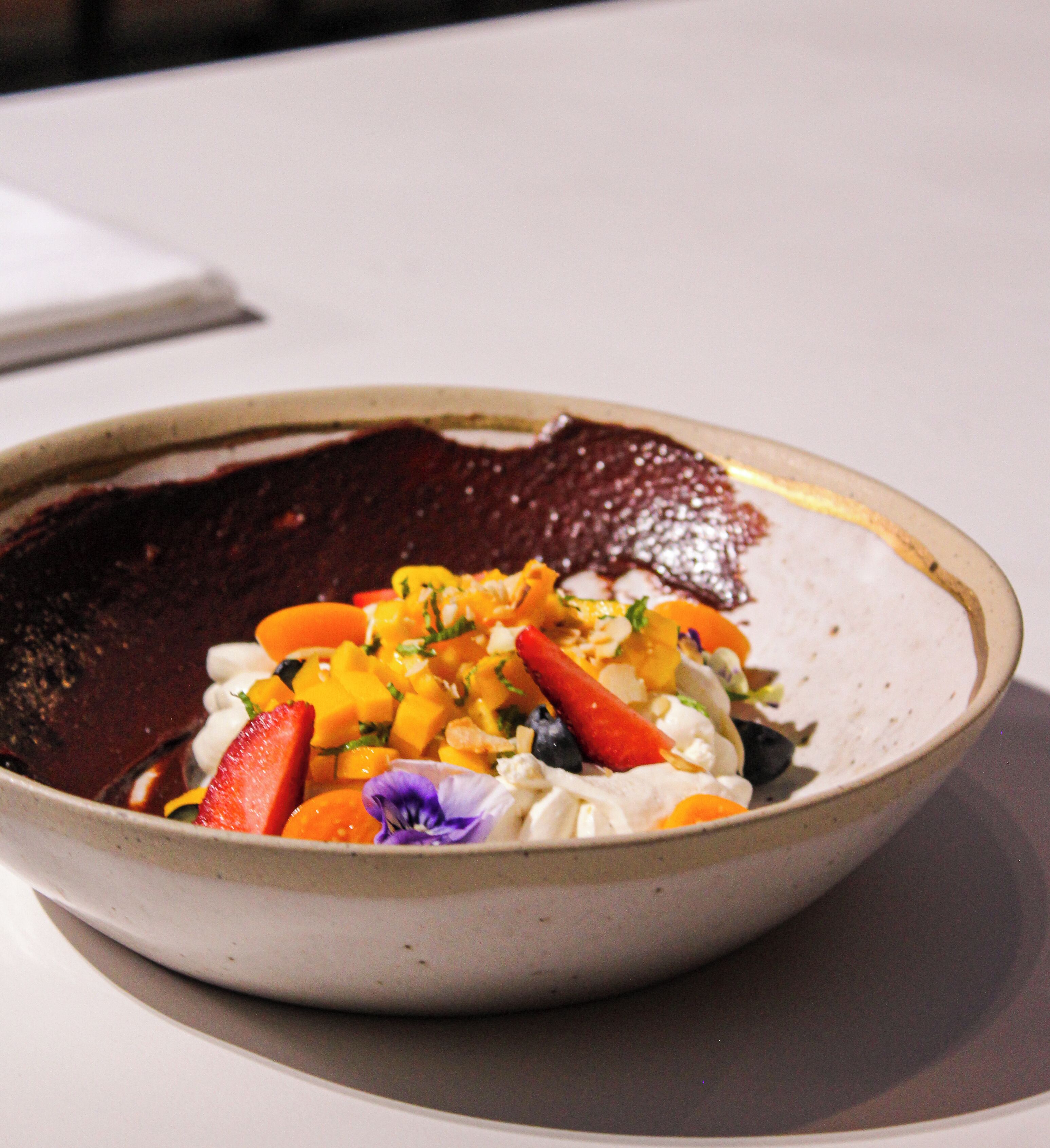A bowl of fruit salad with strawberries, mango, and other fruits on a white table.
