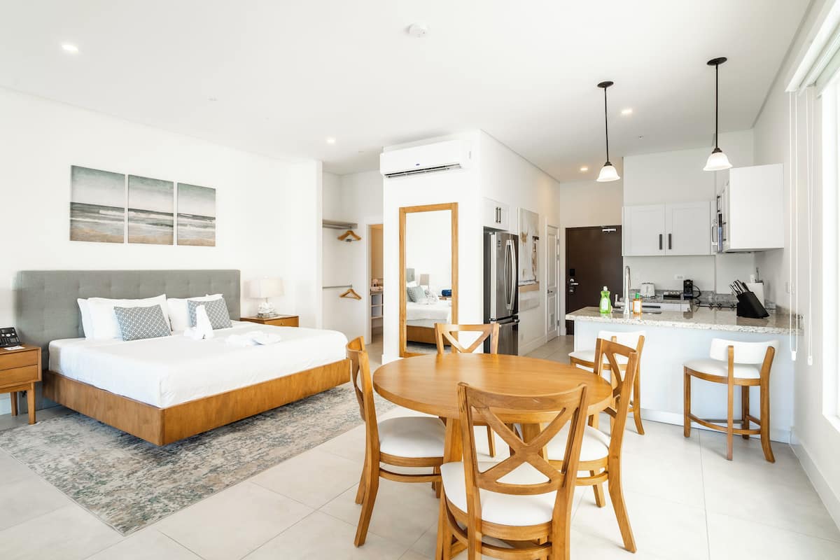 A modern hotel room with a bed, a dining table with chairs, a kitchen area, and a bathroom.