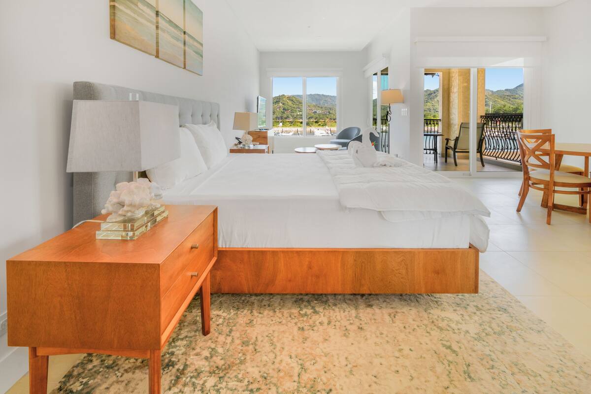 A modern hotel room with a large bed, a wooden nightstand, a desk, and a balcony with a view of the hills.