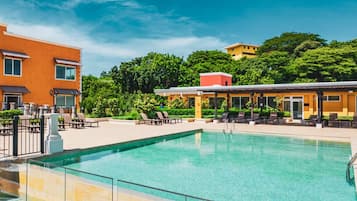A swimming pool with lounge chairs and a covered seating area, surrounded by buildings and greenery.