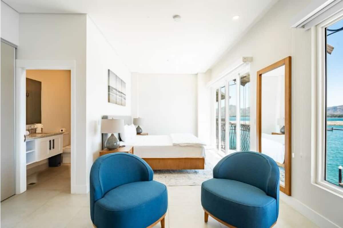 A modern bedroom with a large bed, two blue chairs, a nightstand, and a view of the sea through large windows.