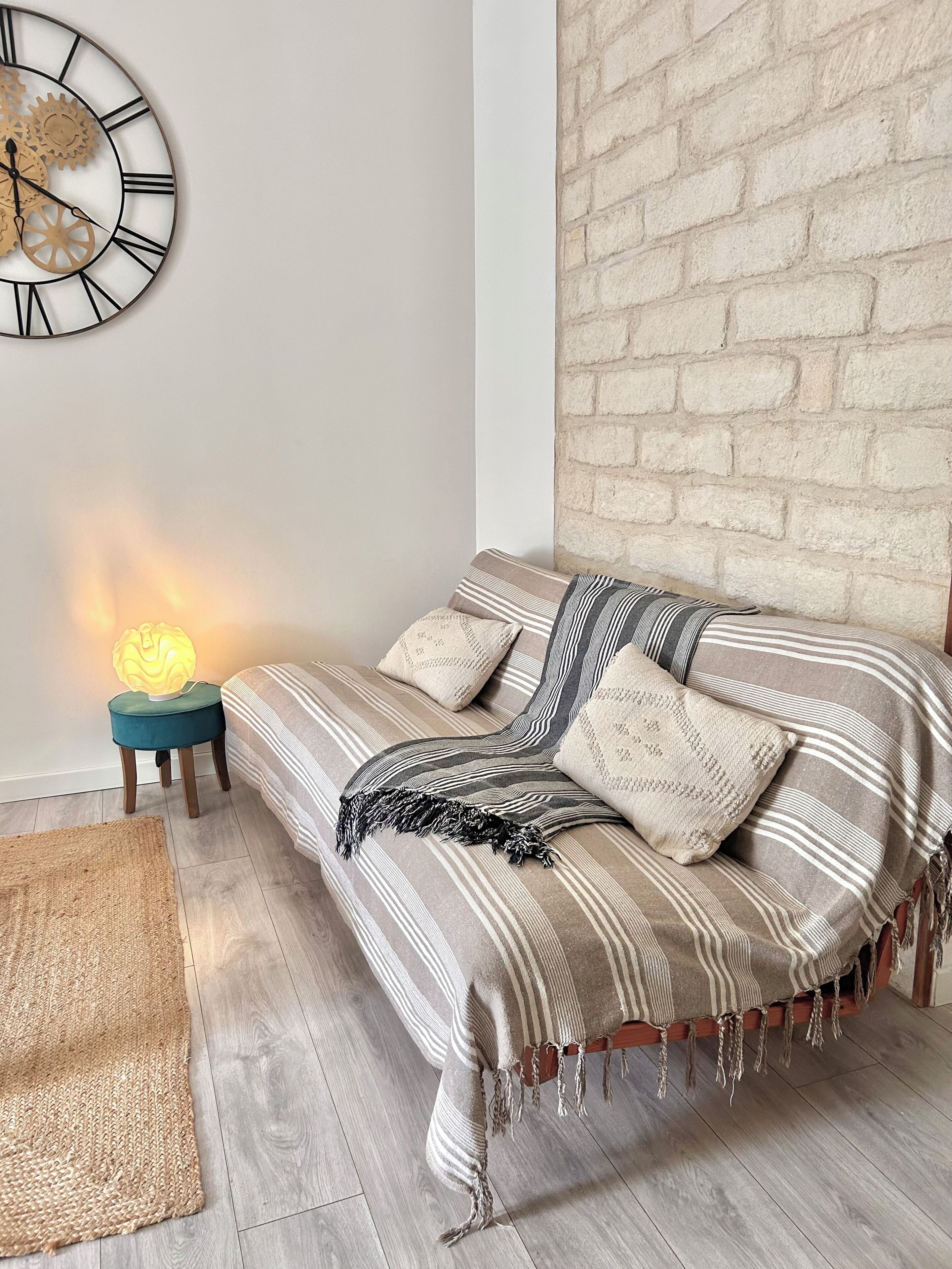 Una habitación con pared de ladrillo, un sofá con ropa de cama a rayas, un reloj con números romanos y una mesita con una lámpara.
