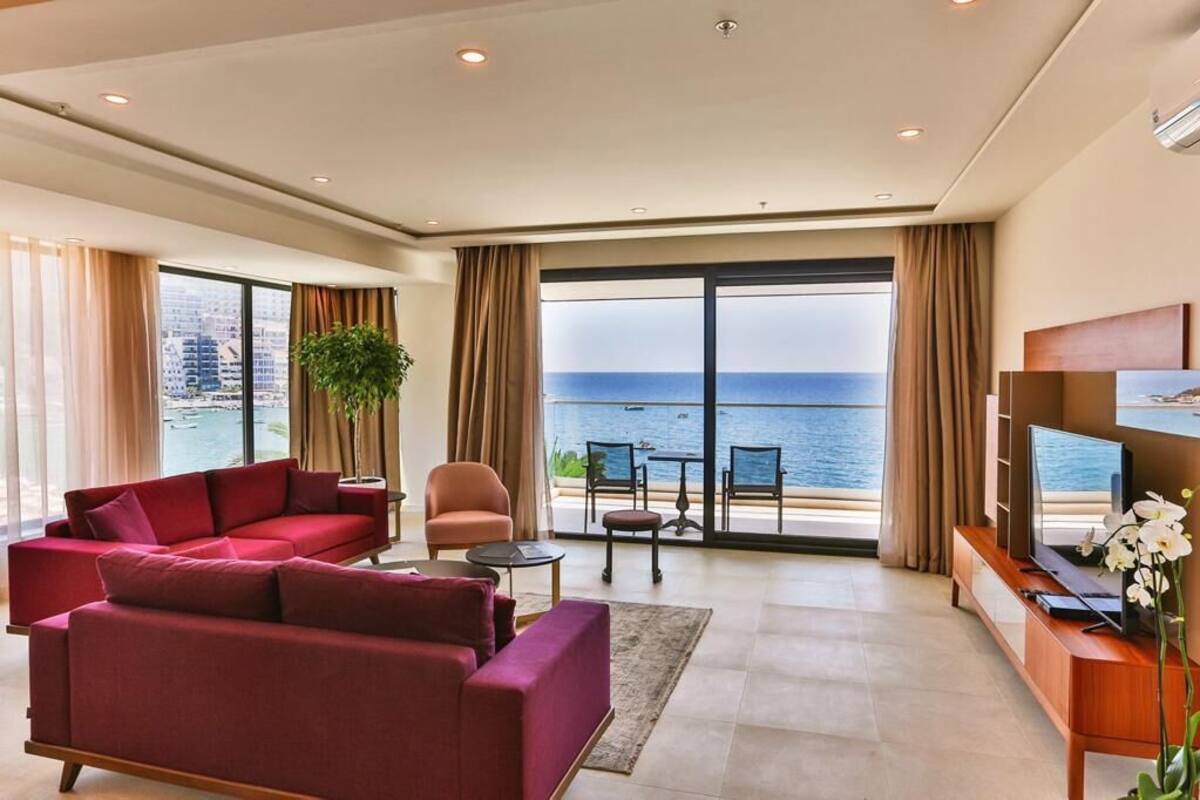 A modern living room with a red sofa, a small table, a TV, and a view of the sea through large windows.