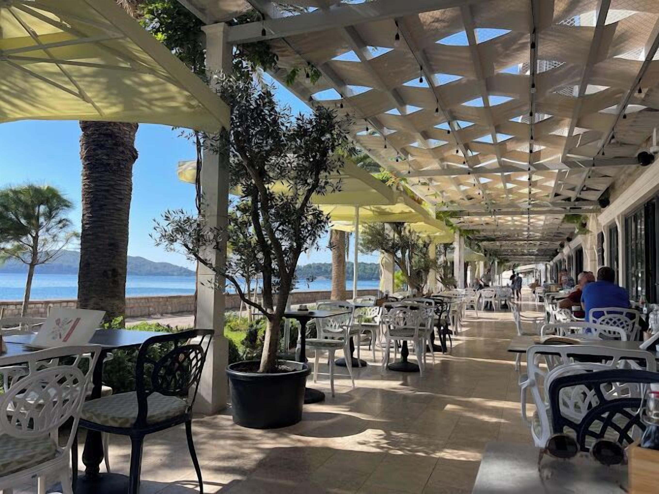 An outdoor cafe with tables and chairs, a view of the sea, and a pergola structure.