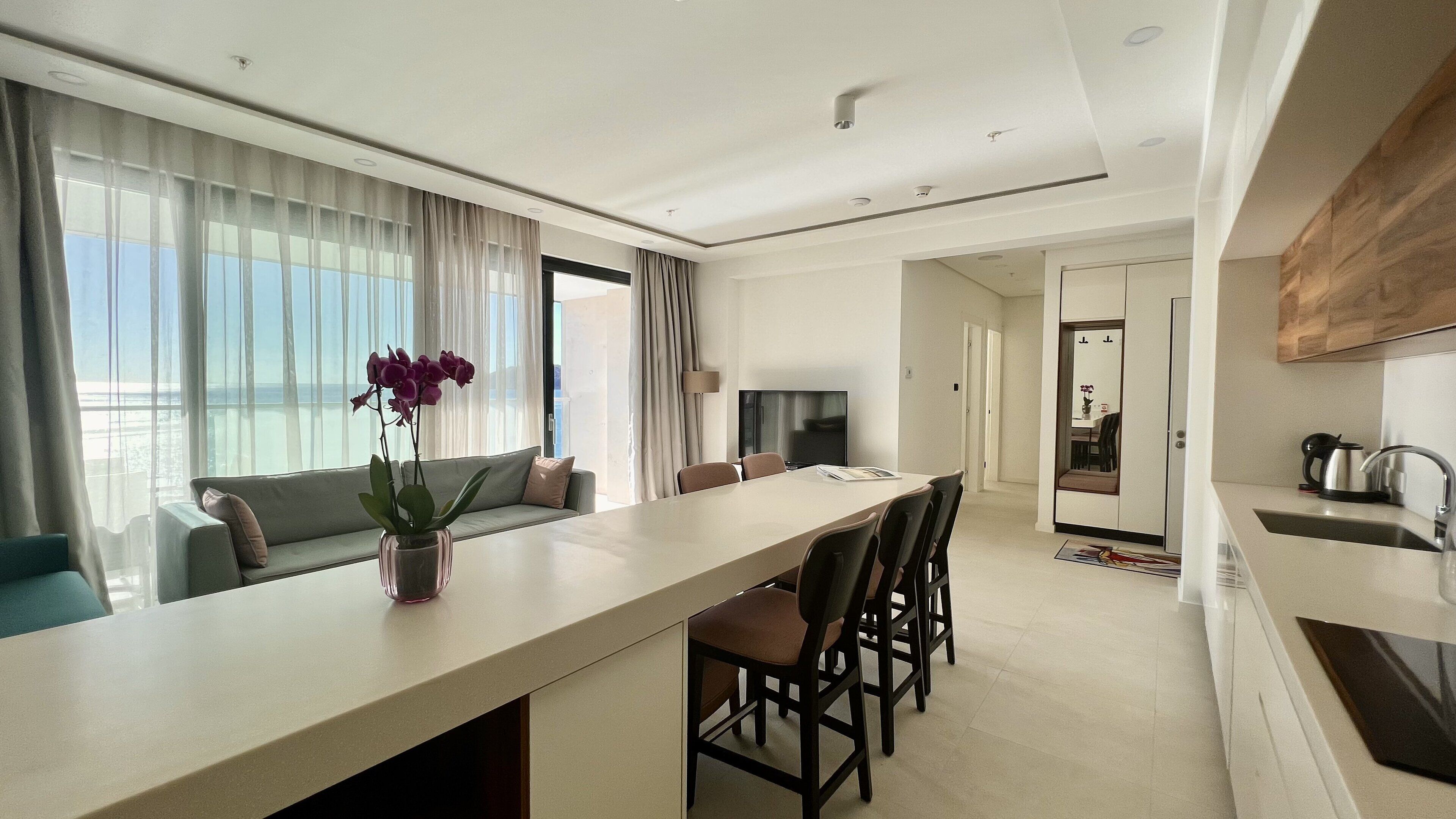 A modern hotel room with a dining area, a sofa, a TV, and a view of the sea.