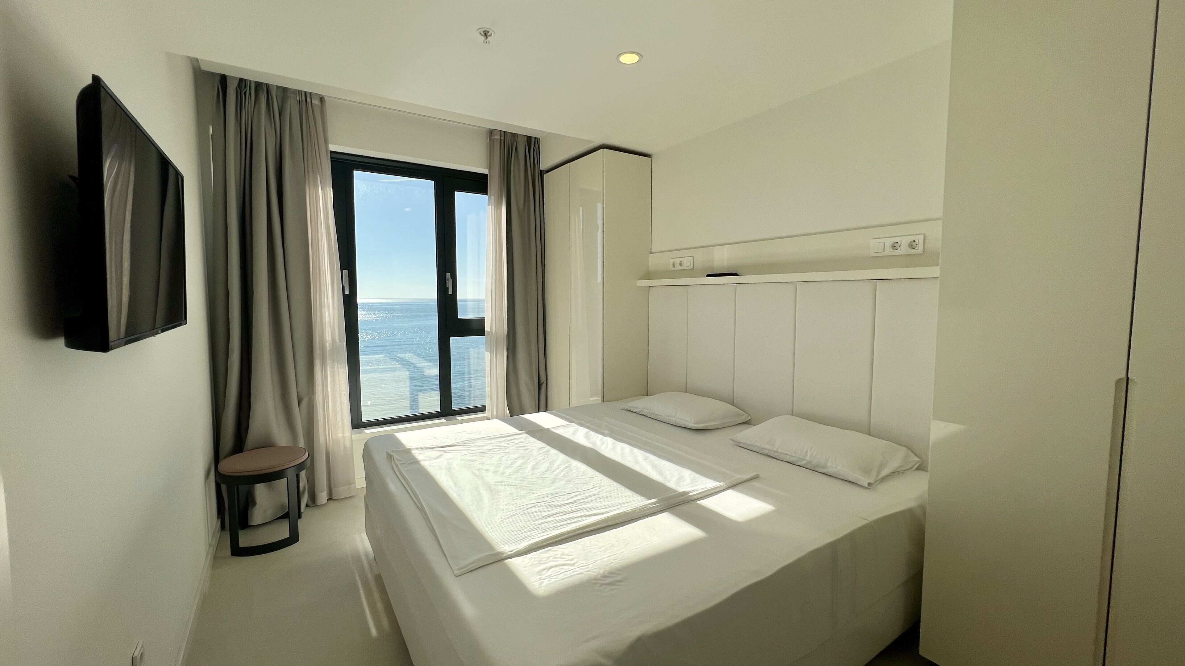 A hotel room with a bed, a TV, a window with curtains, and a view of the sea.