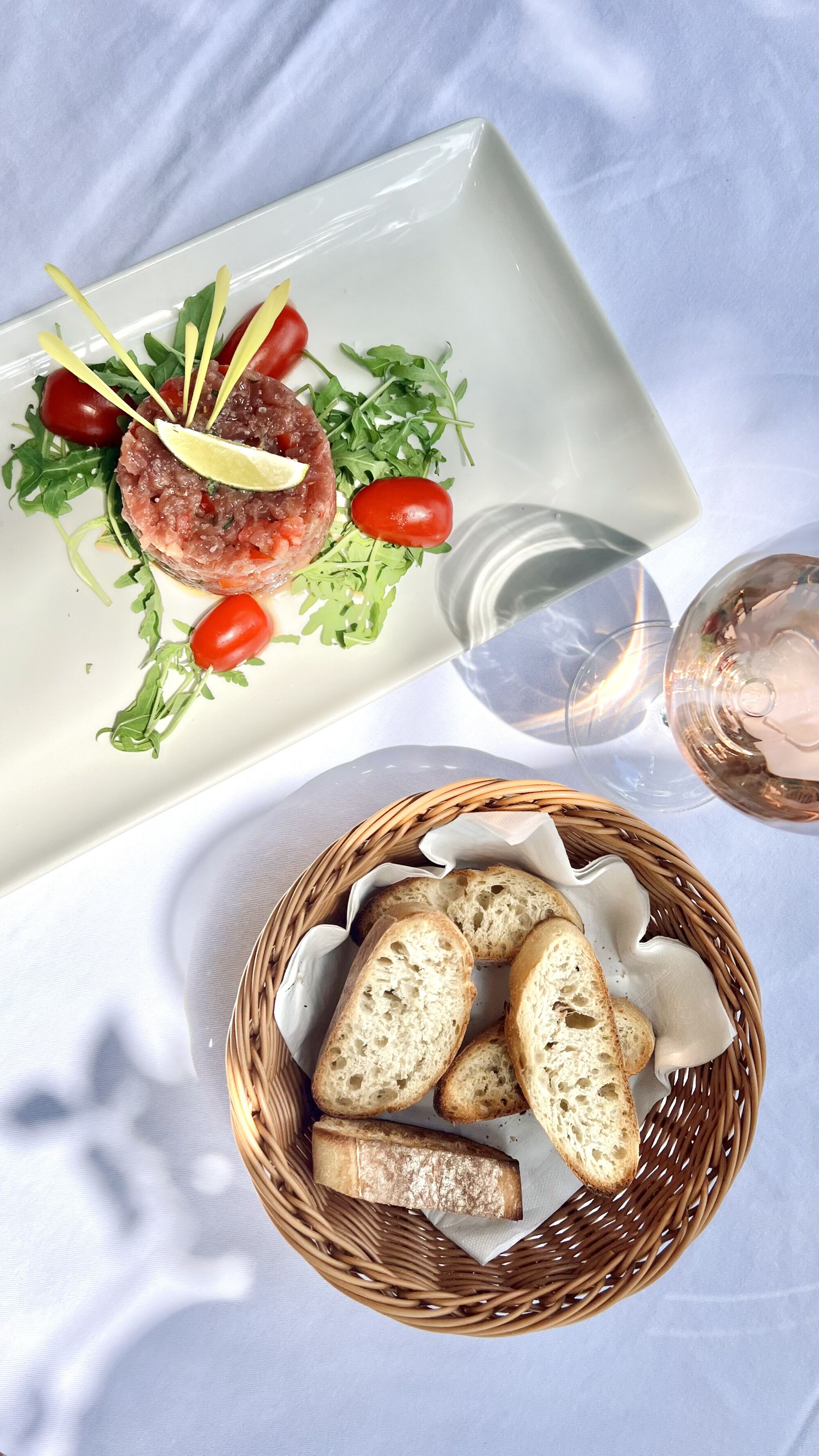 A white plate with a meat dish, cherry tomatoes, and greens, a glass of water, and a basket of bread.
