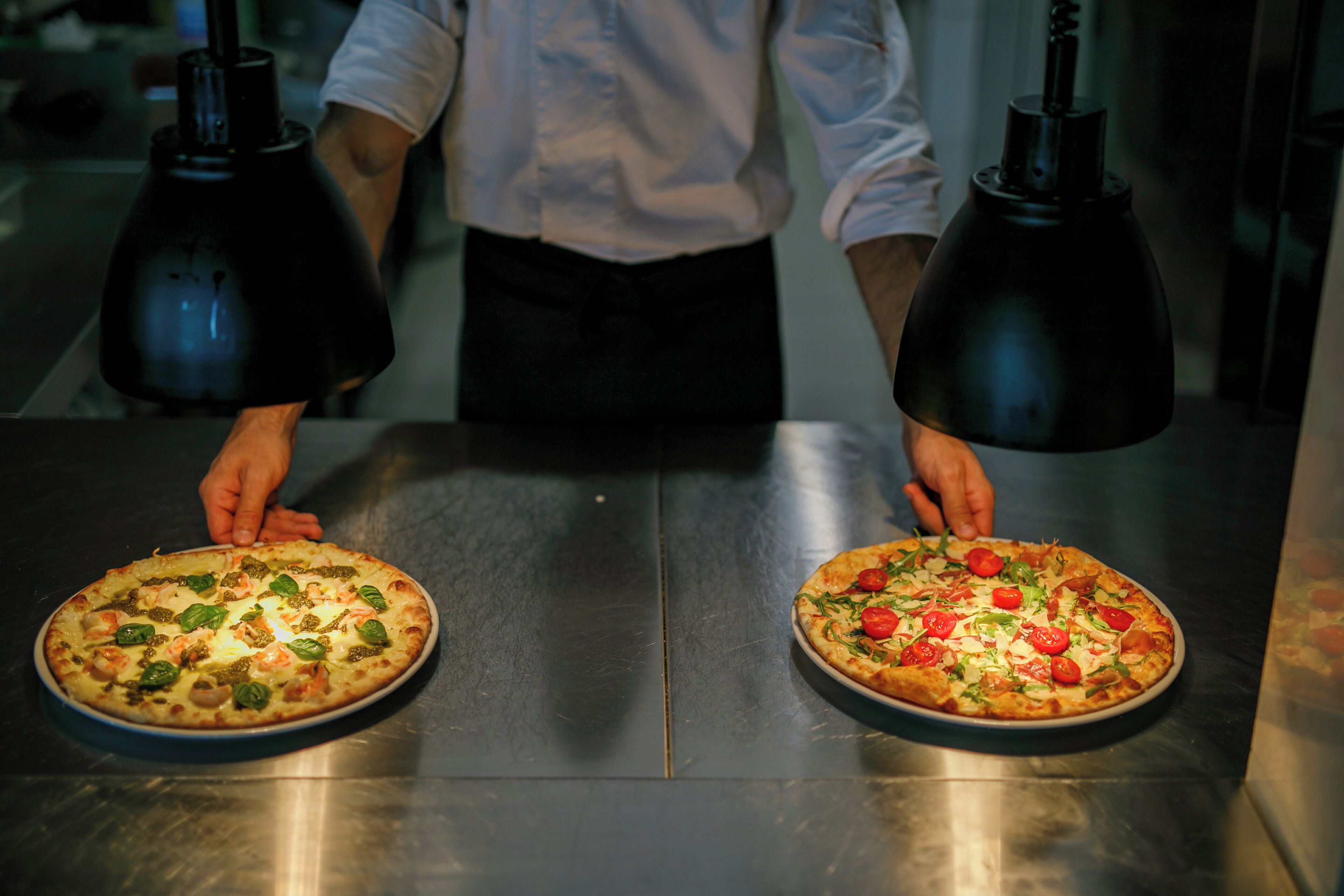 Two pizzas on a stainless steel table with a person standing behind them.