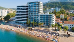 Un hotel moderno con varios pisos y balcones, ubicado cerca de una playa con tumbonas y sombrillas.
