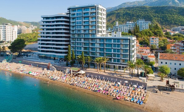 A modern hotel with multiple floors and balconies, situated near a beach with sun loungers and umbrellas.