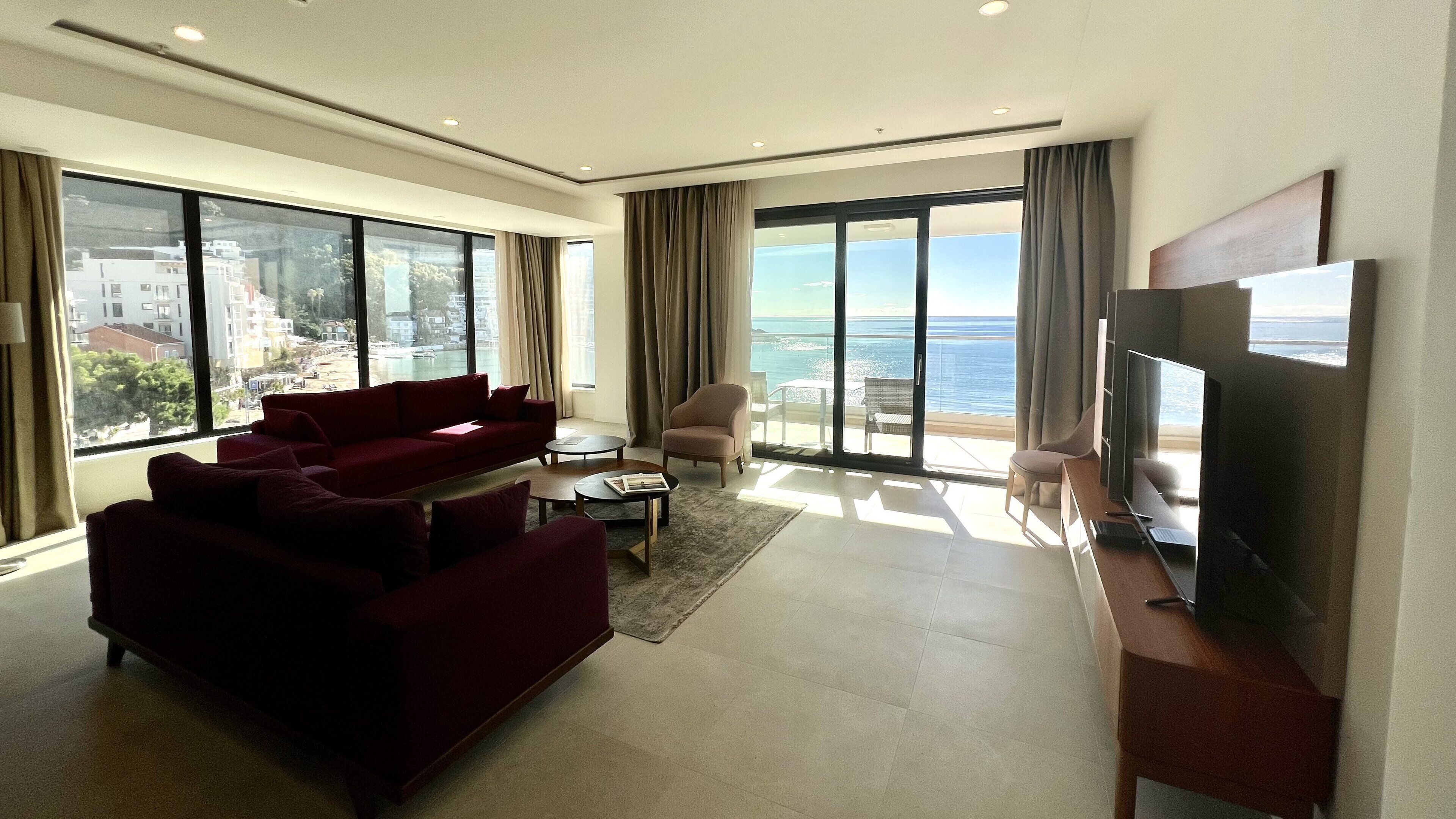 A modern living room with a large sofa, a flat-screen TV, and a balcony with a view of the sea.