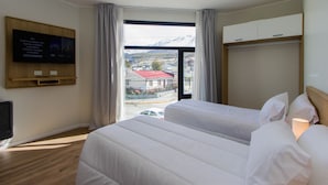 Ein Hotelzimmer mit zwei Betten, einem an der Wand montierten Fernseher, einem Fenster mit Vorhängen und Blick auf eine Stadt und Berge.