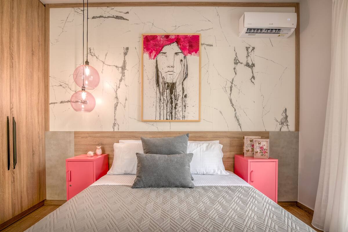 A modern bedroom with a marble-patterned wall, a large bed with patterned bedding, a pink cabinet, and a framed artwork above the bed.