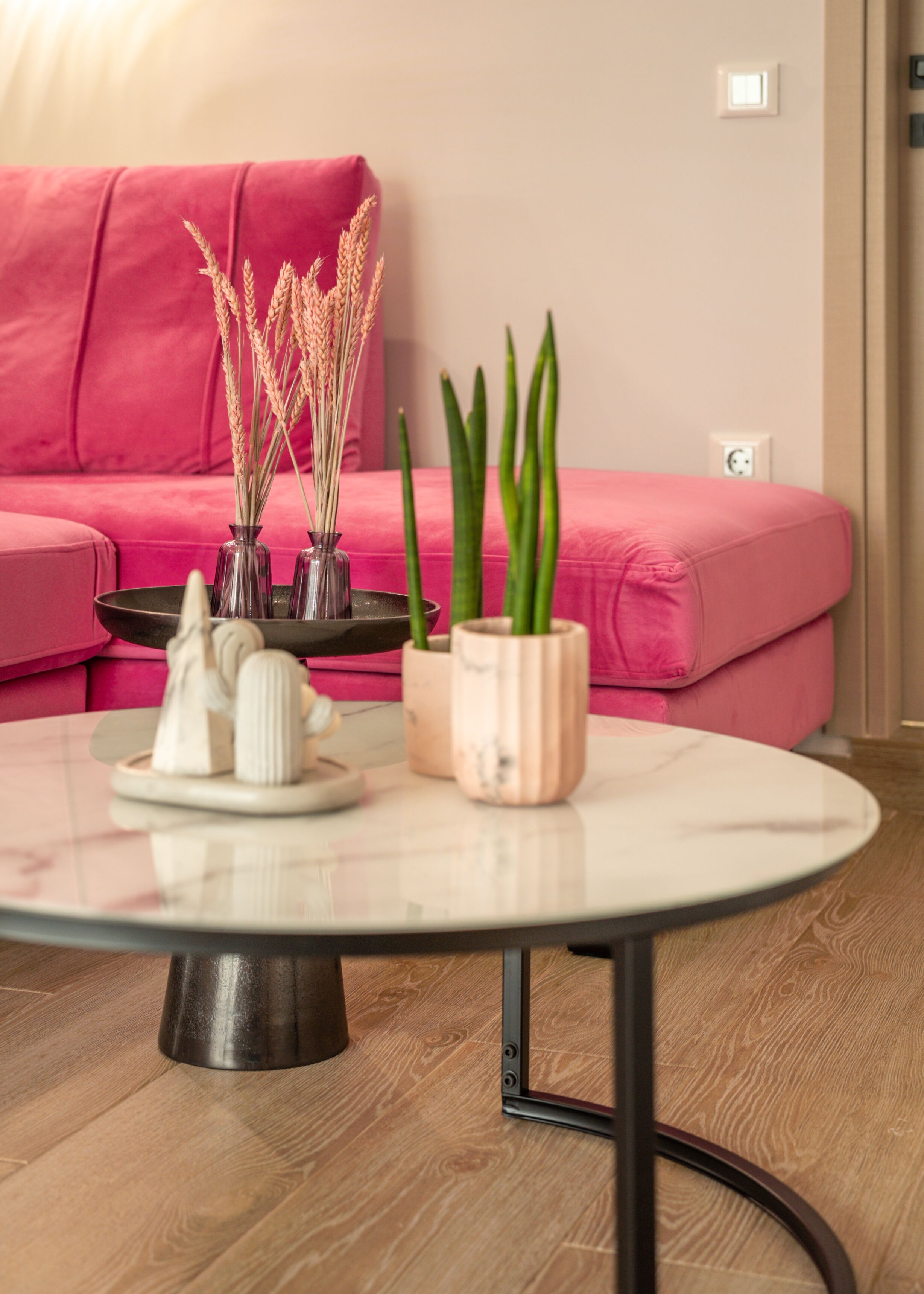 Een ronde glazen tafel met een metalen onderstel, een roze bank en twee potplanten met groene bladeren en lange, dunne stelen.