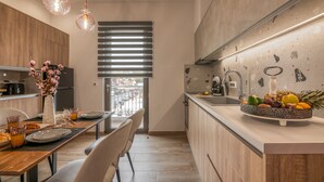 A modern kitchen with wooden cabinets, a dining area with a table set for two, and a fruit bowl on the counter. - MAGNOLIA Luxury Chania Central Apartments (Chania)