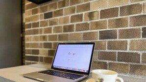 Un ordinateur portable est posé sur une table, avec une tasse à côté, le tout sur fond de mur en briques.