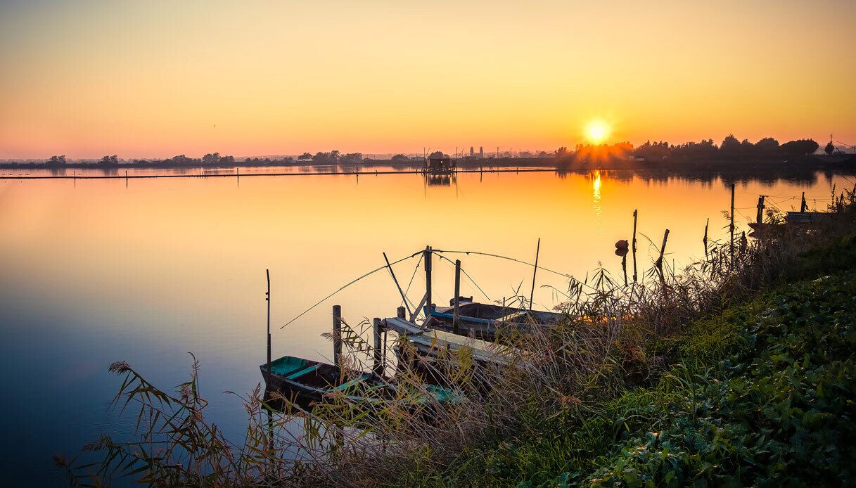 Ein ruhiger See bei Sonnenaufgang, mit Booten, die an der Küste liegen.