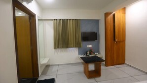 Ein kleiner, sauberer Raum mit gefliestem Boden, einer Holztür, einem kleinen Tisch, einem an der Wand montierten Fernseher und einem Fenster mit Vorhängen.