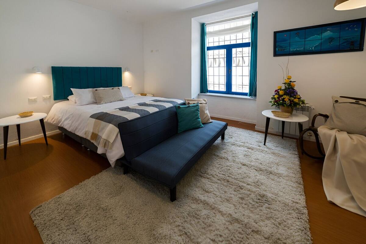 A modern bedroom with a bed, bench, bedside tables, a window with curtains, a small table with a plant, and a chair.