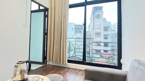 A modern living room with a sofa, a small table with a kettle and a tray, a window with curtains, and a view of buildings outside. - Hanowood Homestay & Travel (Hanoi)