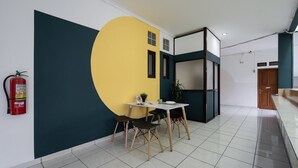 A modern interior with a dining area featuring a white table and chairs, a yellow accent wall, and a fire extinguisher mounted on the wall. - Cove Horizone (Bandung)