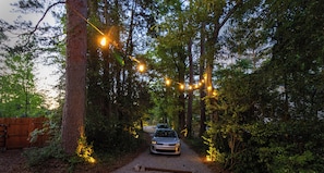 A gravel path leading through a wooded area with string lights overhead and a car parked at the end.