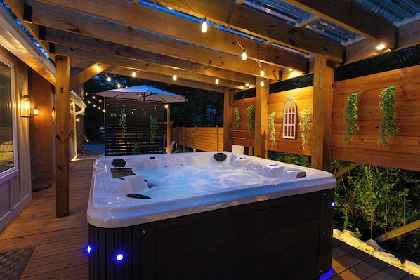 A hot tub under a wooden pergola with string lights, surrounded by wooden walls and greenery.
