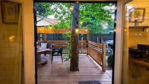 Eine Holzterrasse mit Sitzgelegenheiten, ein Baum und Blick auf einen Garten mit Zaun und Fahrzeugen.
