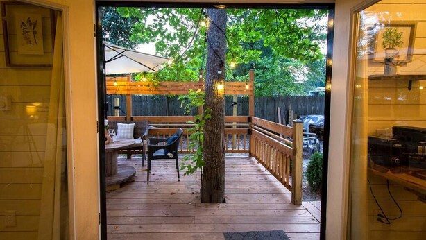 A wooden deck with outdoor seating, a tree, and a view of a backyard with a fence and vehicles.