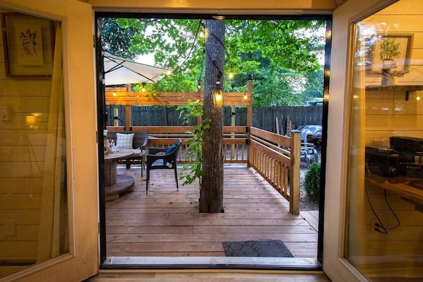 Une terrasse en bois avec des sièges d’extérieur, un arbre et une vue sur une cour arrière avec une clôture et des véhicules.