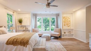 Ein Schlafzimmer mit einem großen Bett, einem Deckenventilator, einem Fenster mit Vorhängen, einem kleinen Tisch mit einer Pflanze und einem Wanddekor.