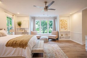 Une chambre avec un grand lit, un ventilateur de plafond, une fenêtre avec des rideaux, une petite table avec une plante et un élément décoratif mural.
