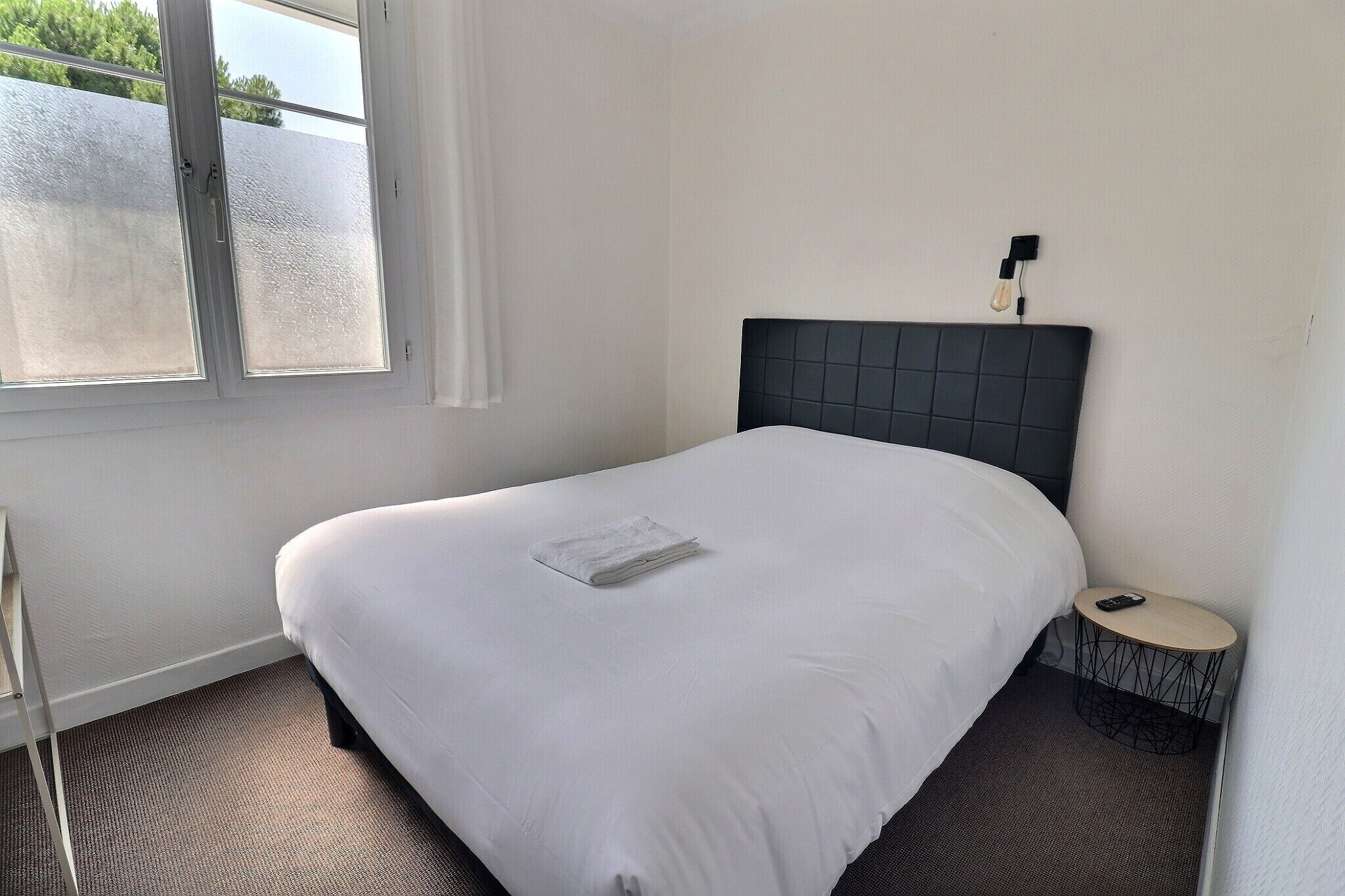 A neatly made bed with white linens, a small bedside table with a lamp, and a window with frosted glass.
