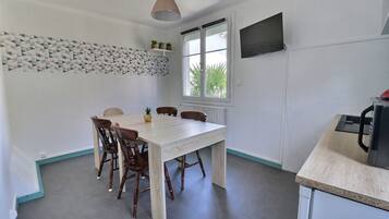A dining area with a white table and wooden chairs, a hanging light fixture, a window with a view of greenery, and a kitchen area with a microwave.