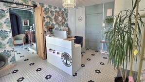 Un área de recepción con un escritorio blanco, una araña, una planta grande en maceta y una pared con estampado tropical.