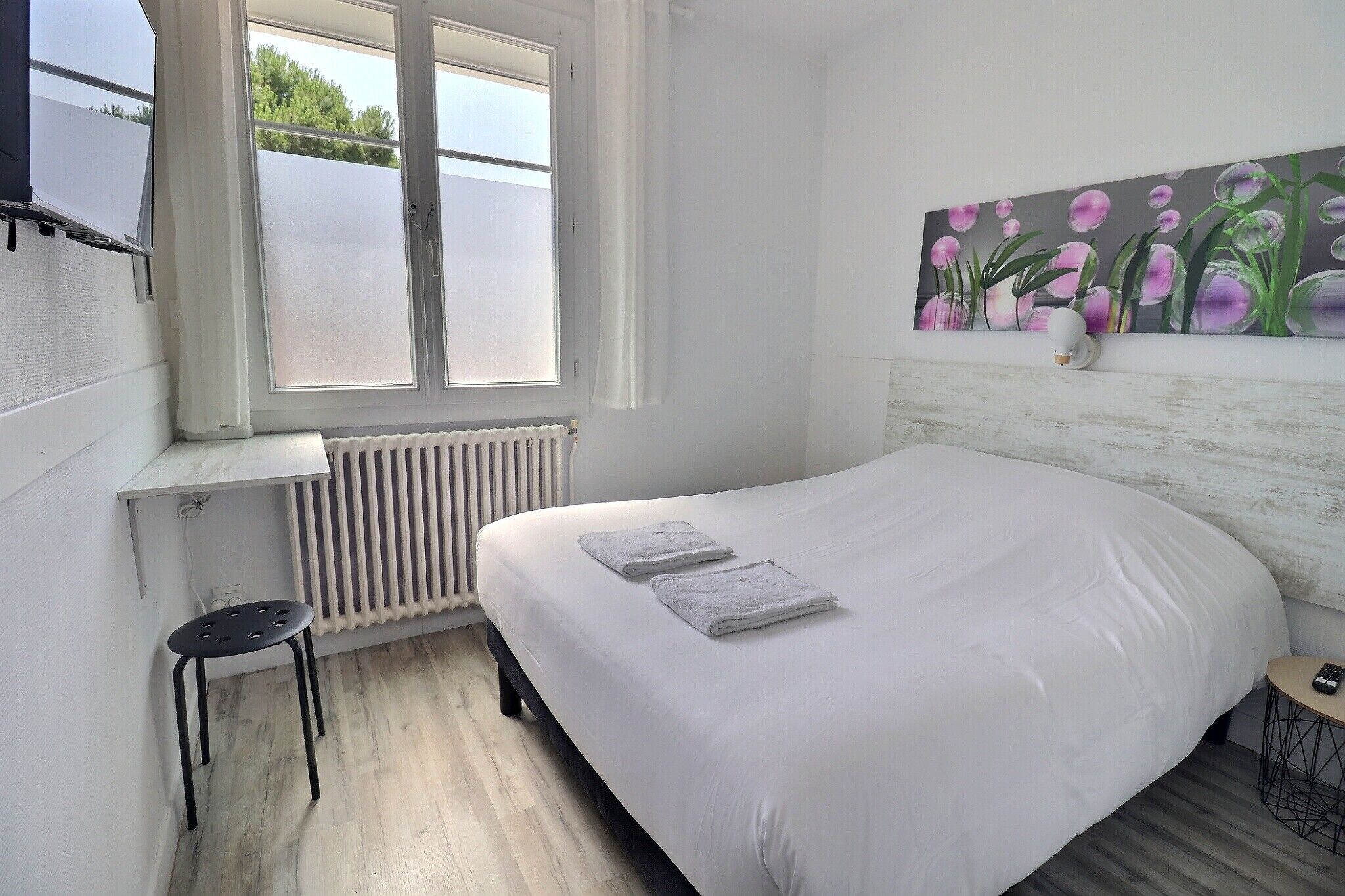 Een slaapkamer met een bed, een raam, een radiator, een klein krukje en een schilderij aan de muur.