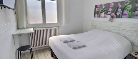 A bedroom with a bed, a window, a radiator, a small stool, and a painting on the wall.