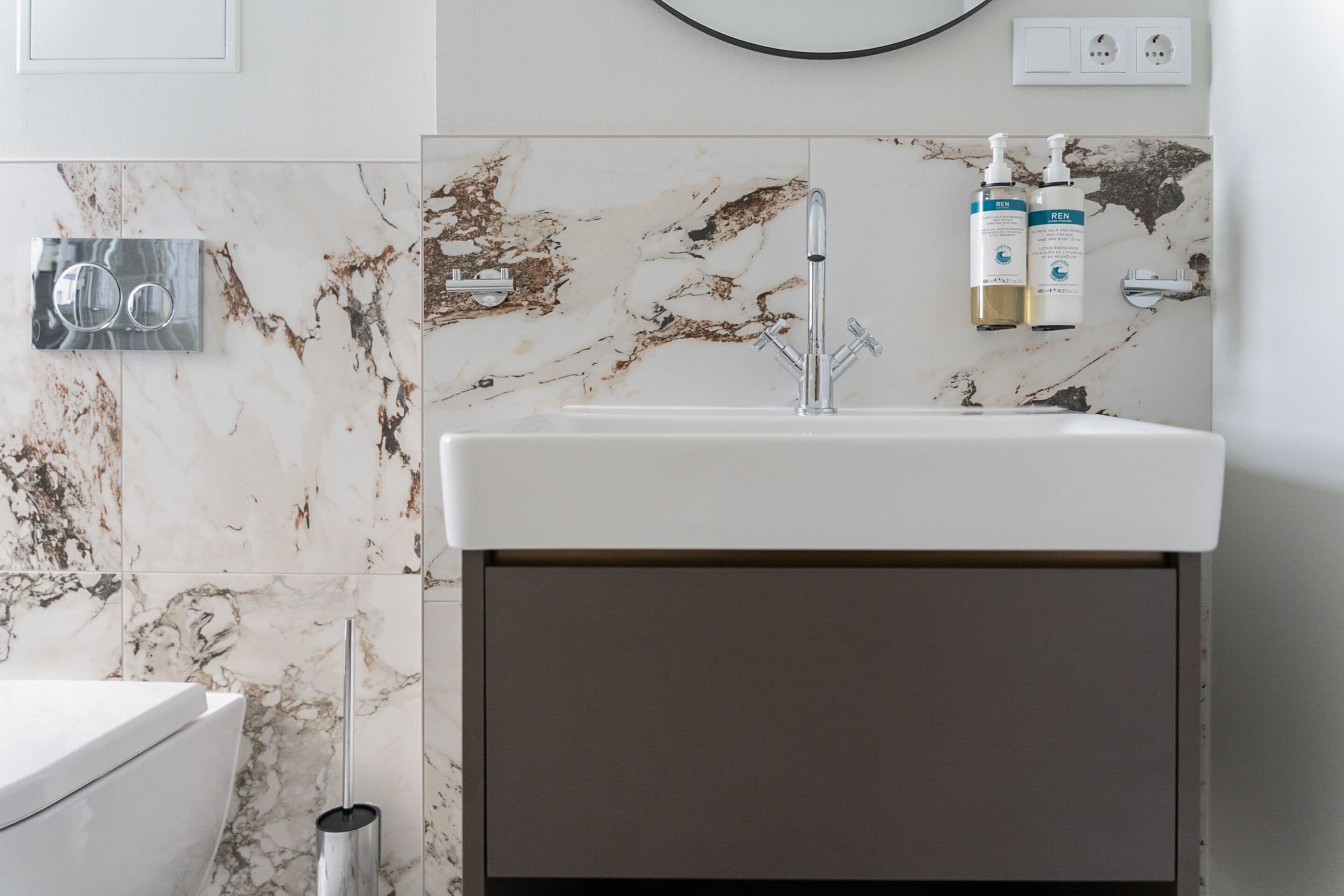 Een badkamer met een witte wasbak, een marmeren achterwand en twee flessen REN shampoo en conditioner.
