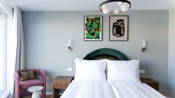 Un dormitorio moderno con una cama grande, una mesita redonda pequeña, un sillón morado y dos lámparas fijadas a la pared.